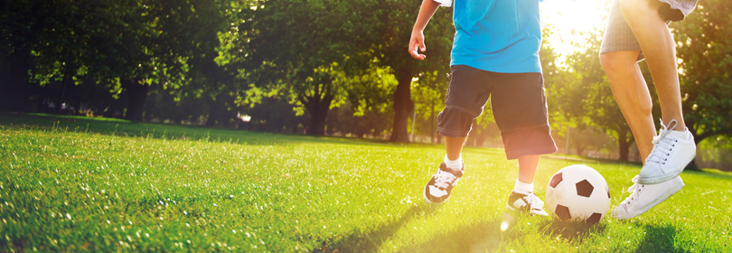 Two children kicking a soccer ball in a field of grass with trees in the background